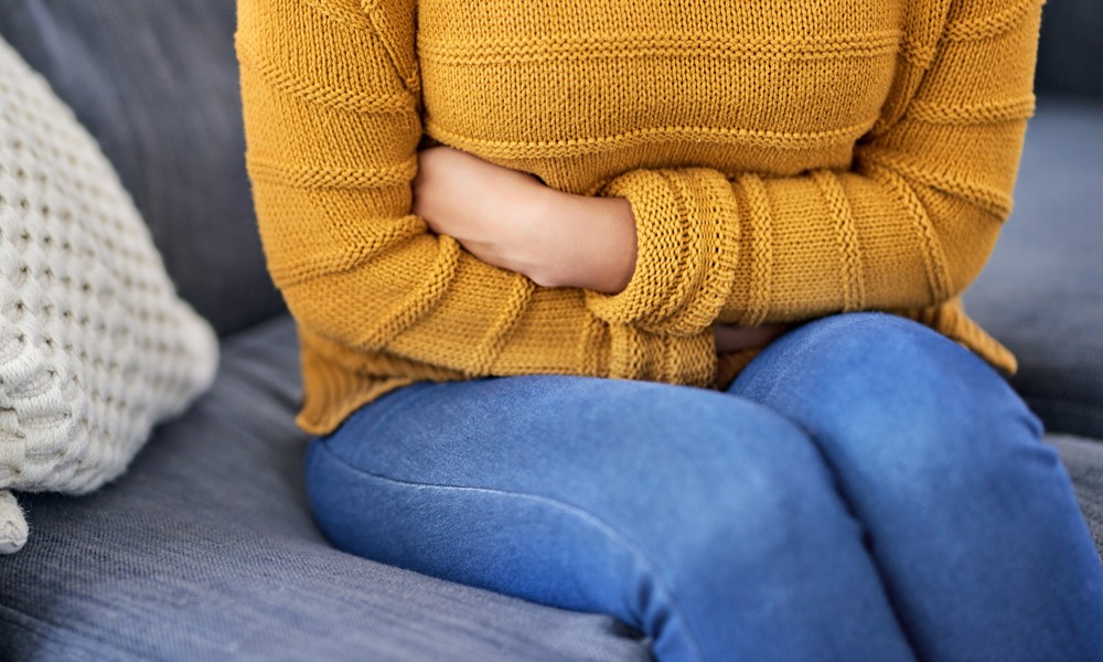 Frau in blauer Jeans und gelben Oberteil sitzt schmerzerfüllt auf einer Couch 